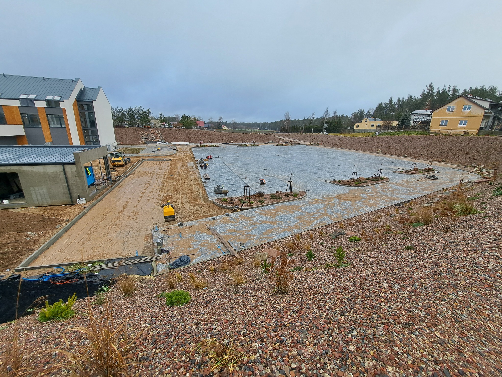 Układanie kostki brukowej na dużej powierzchni z elementami zieleni. Widoczny budynek w tle i przygotowane podłoże. Prace brukarskie w toku, z użyciem zagęszczarki.