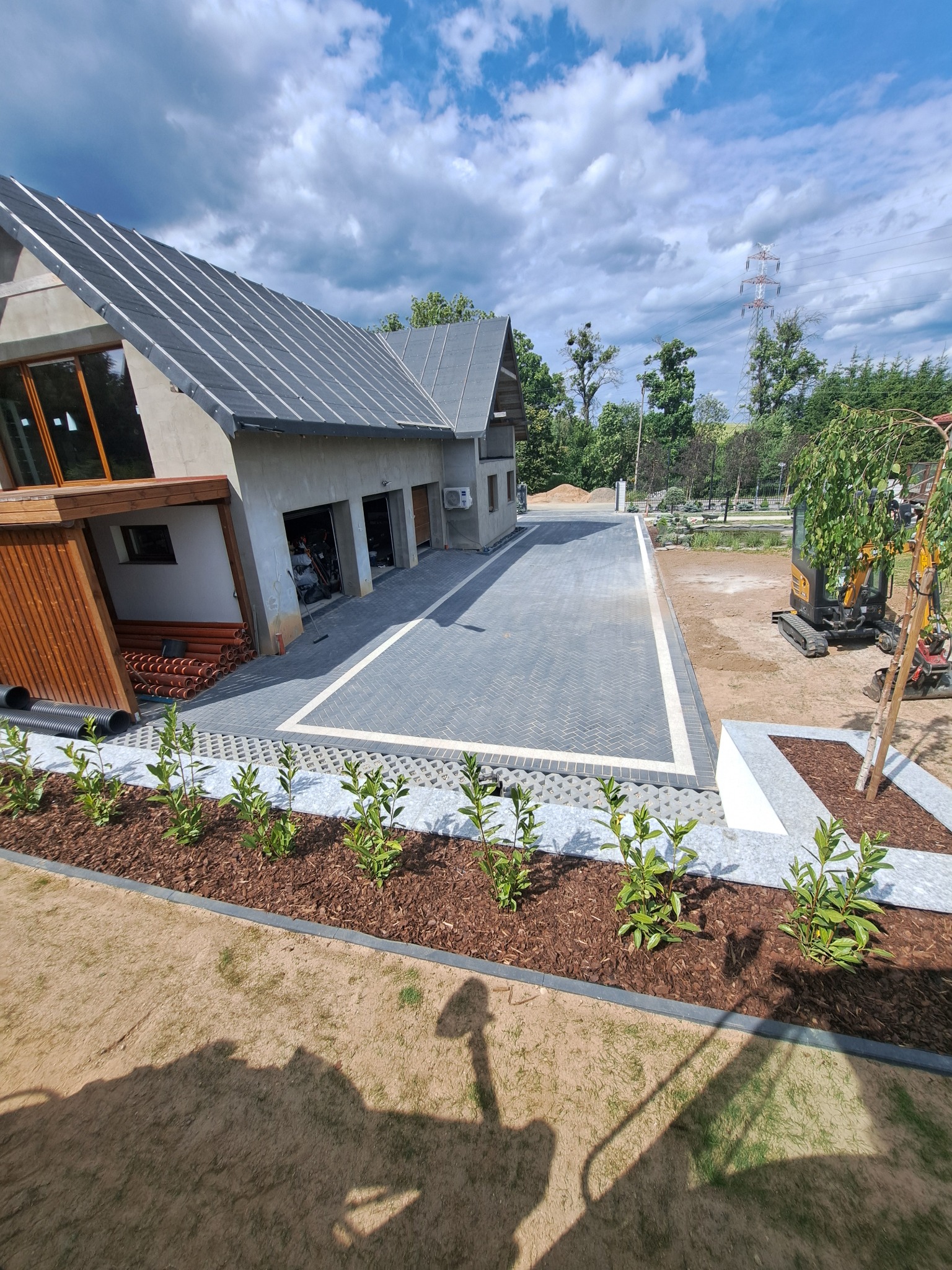 Nowoczesny dom z szarej kostki brukowej ułożonej w jodełkę, obrzeża z jasnej kostki. Rabaty z korą i zielenią. W tle budynek z garażami i fragment ogrodu z mini koparką.