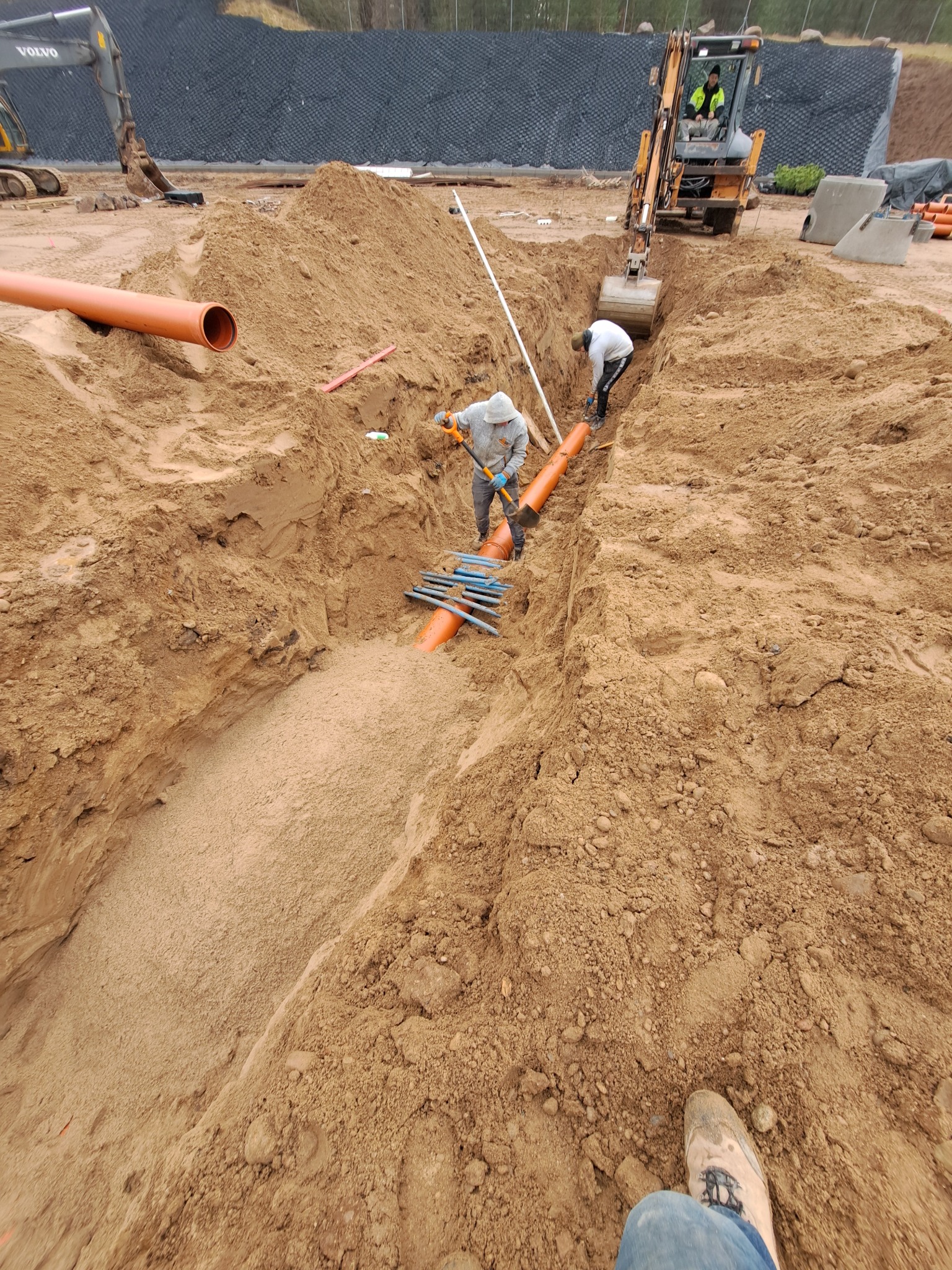 Montaż pomarańczowych rur w wykopie przez dwóch pracowników, z koparką w tle. Widoczne elementy złączek i narzędzia. Prace ziemne w toku, teren budowy.