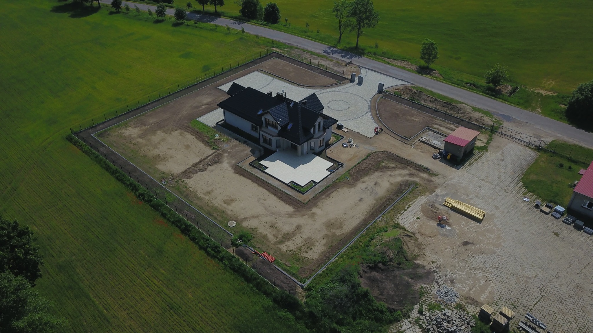 Widok z lotu ptaka na dom z czarnym dachem i jasną elewacją, otoczony terenem w trakcie zagospodarowania, z widoczną kostką brukową i ogrodzeniem.