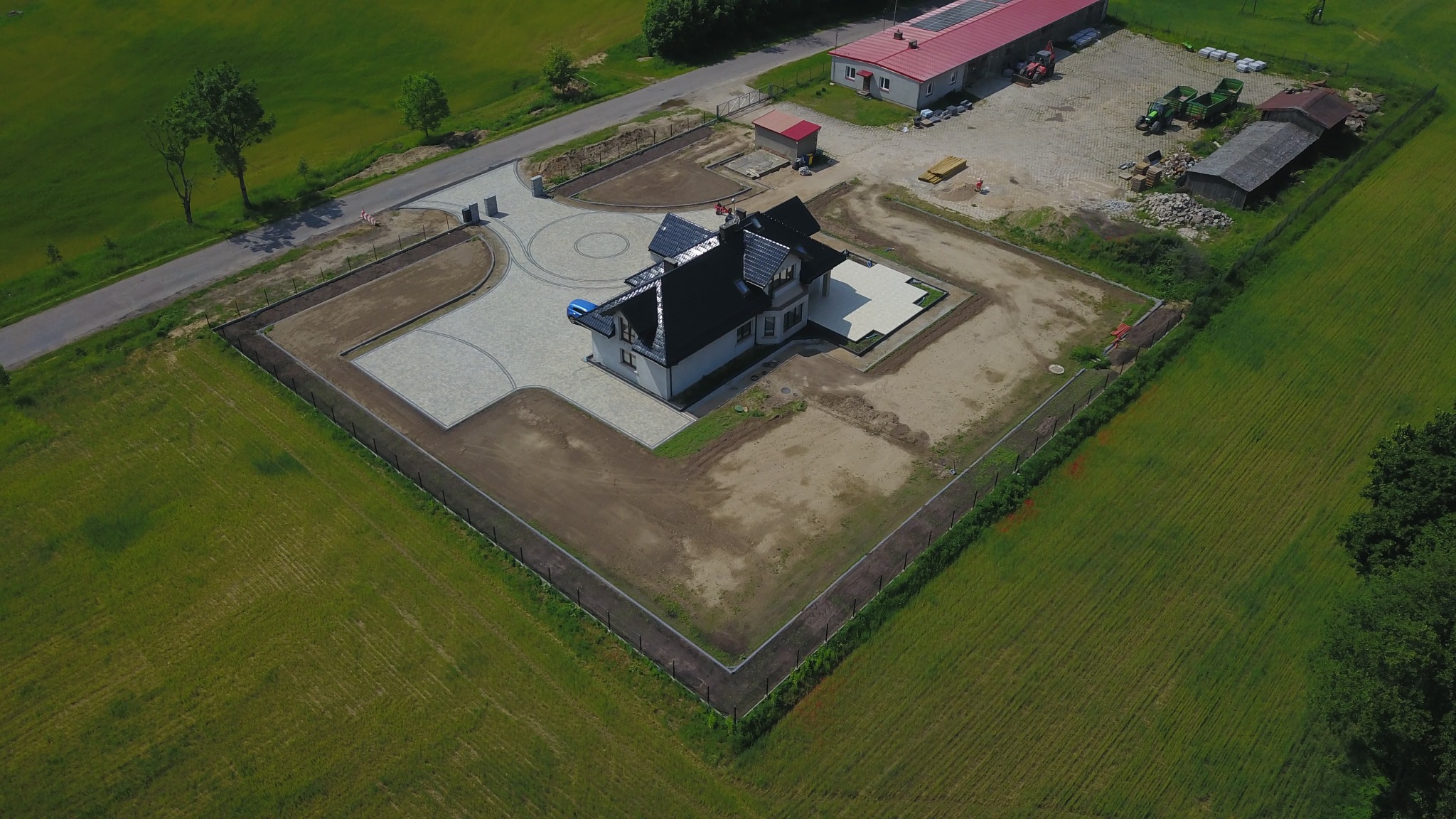 Widok z drona na dom z czarnym dachem i ułożoną kostką brukową na podjeździe. W tle budynki gospodarcze i pola uprawne. Całość otoczona ogrodzeniem.