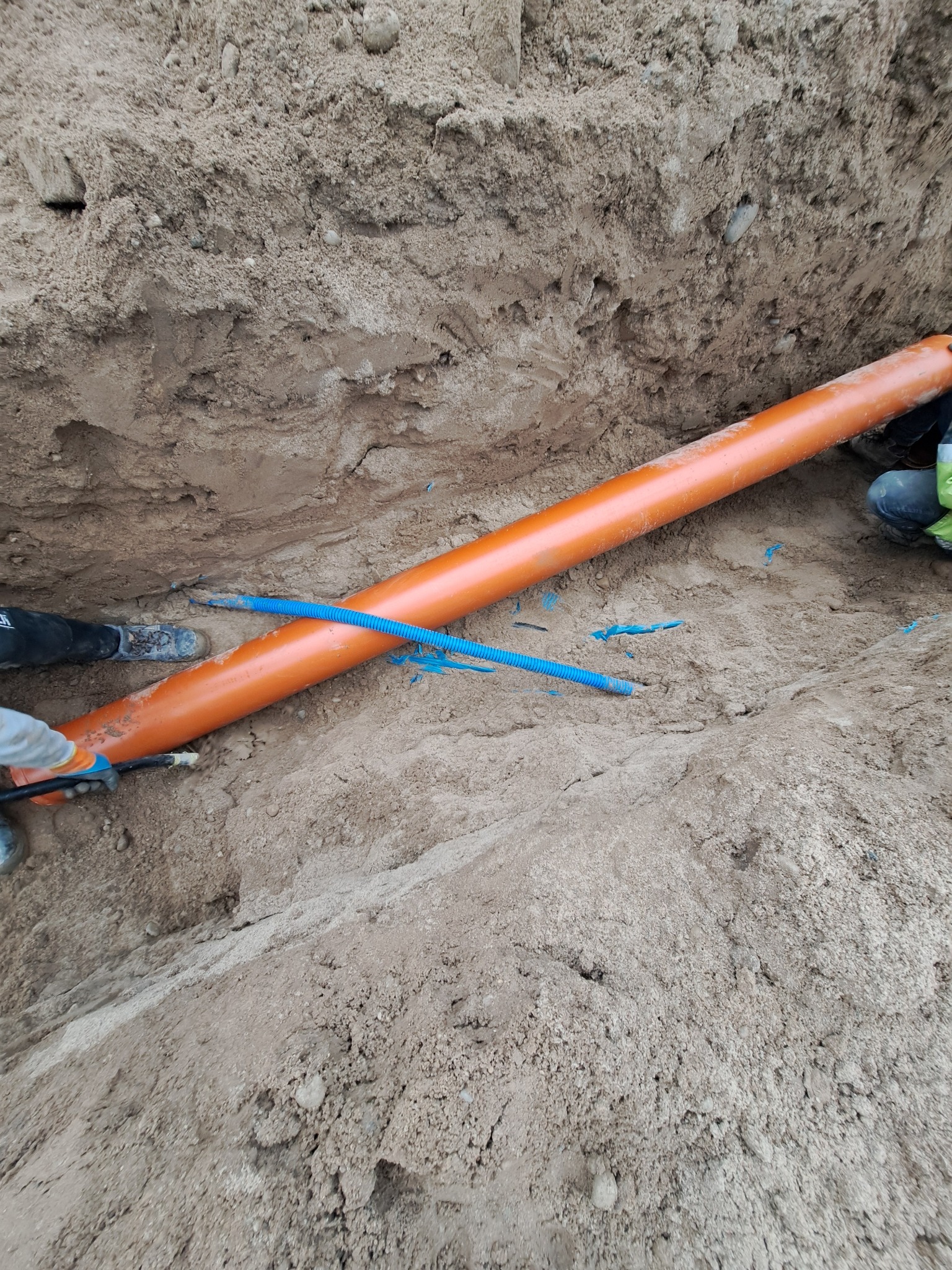 Pomarańczowa rura kanalizacyjna w wykopie ziemnym, częściowo zasypana piaskiem. Widoczny fragment niebieskiej rury karbowanej i fragmenty odzieży pracowników.