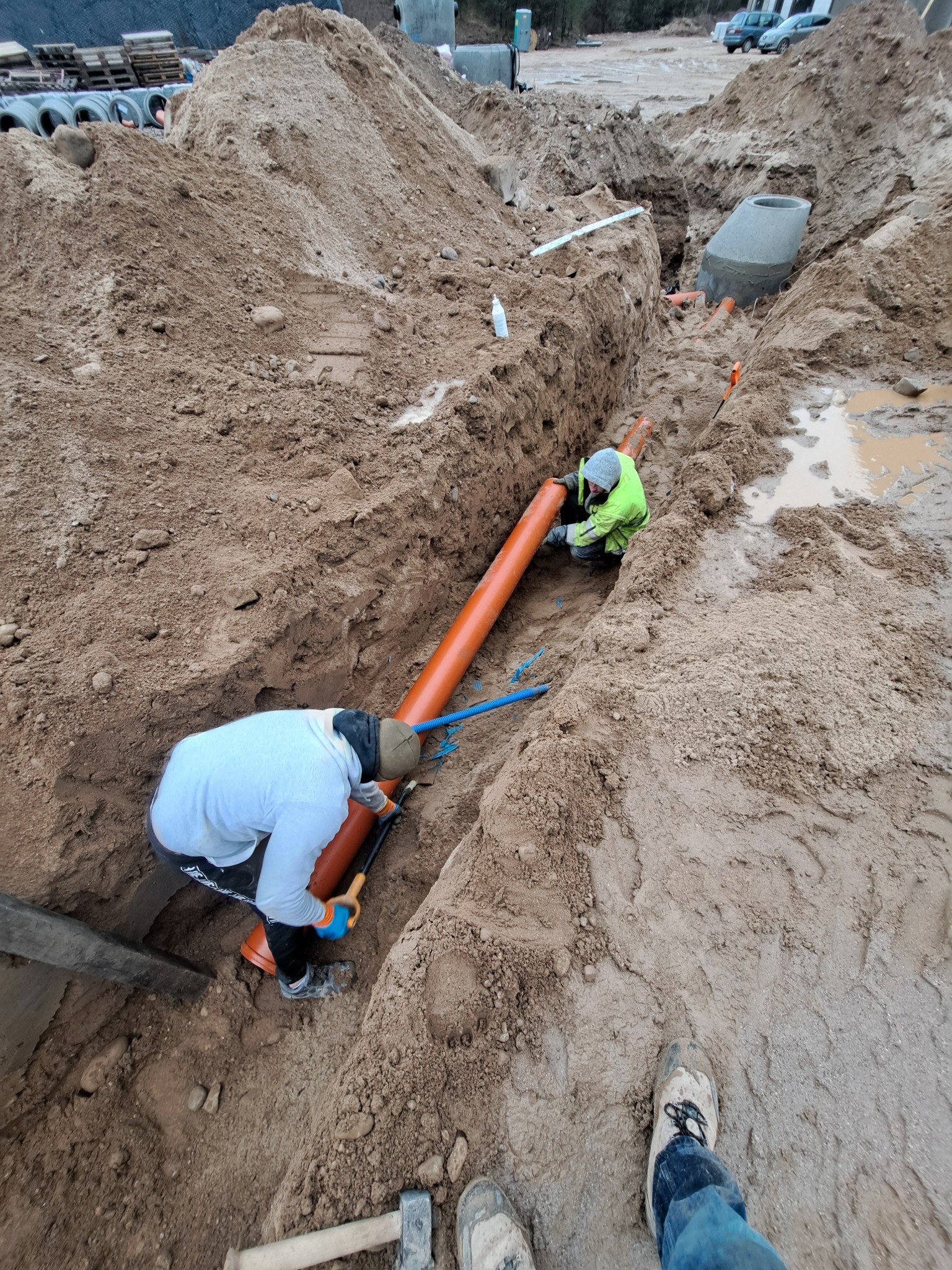 Montaż pomarańczowej rury w wykopie przez dwóch pracowników w odzieży roboczej. W tle widoczny betonowy element kanalizacyjny i hałda ziemi.