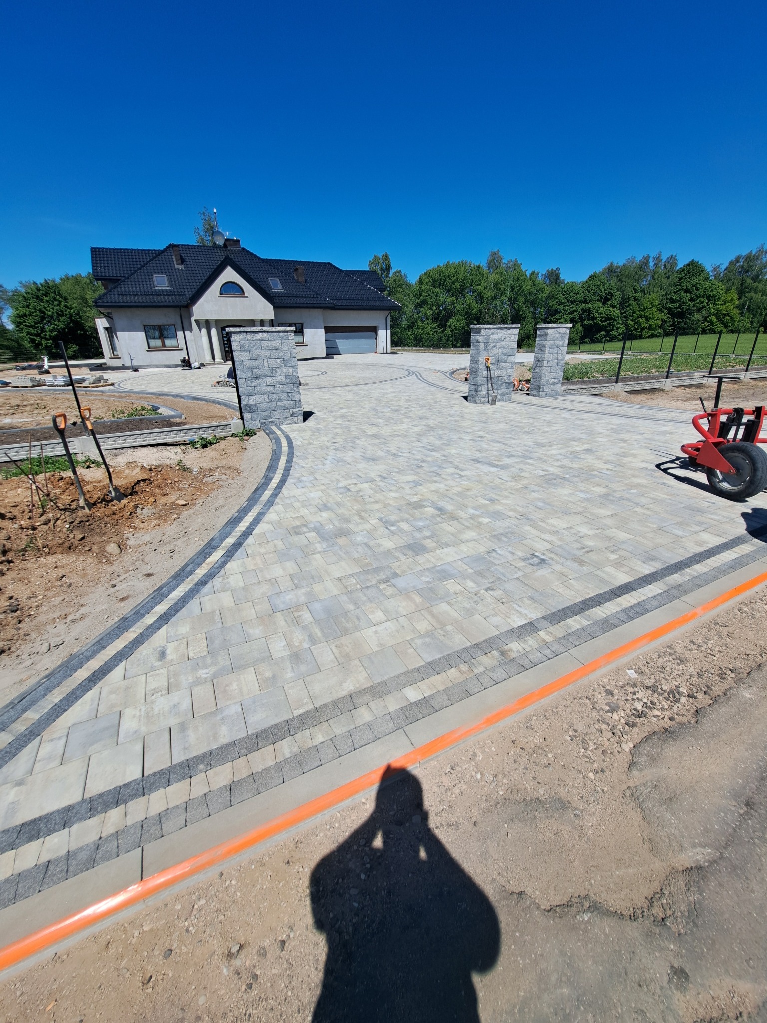 Ułożona kostka brukowa na podjeździe do domu z szarej kostki z ciemnymi obramowaniami. Widoczne słupki z kostki po bokach wjazdu i fragment domu w tle.