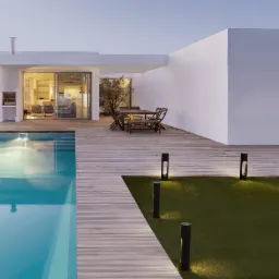 Nowoczesny dom z basenem oświetlony lampami ogrodowymi na trawniku i drewnianym tarasie. Minimalistyczny design, ciepłe światło wieczorne.