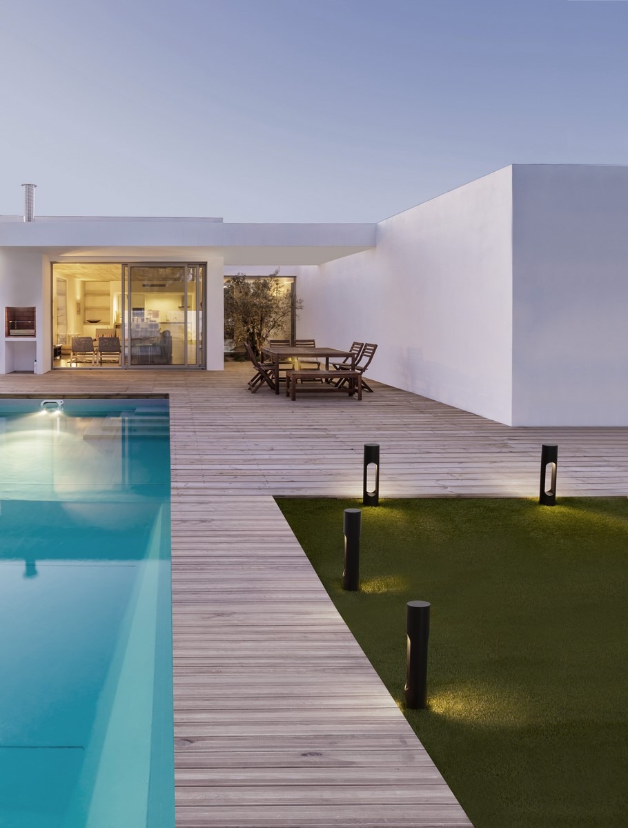 Nowoczesny dom z basenem oświetlony lampami ogrodowymi na trawniku i drewnianym tarasie. Minimalistyczny design, ciepłe światło wieczorne.