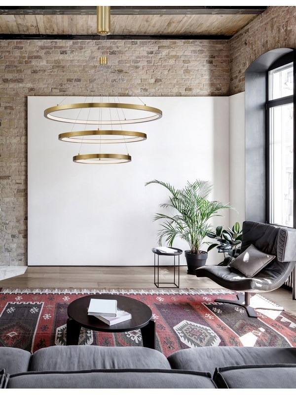 Nowoczesna aranżacja wnętrza z trzema okrągłymi lampami LED w złotym kolorze, zawieszonymi nad dywanem z etnicznym wzorem i szarą sofą.