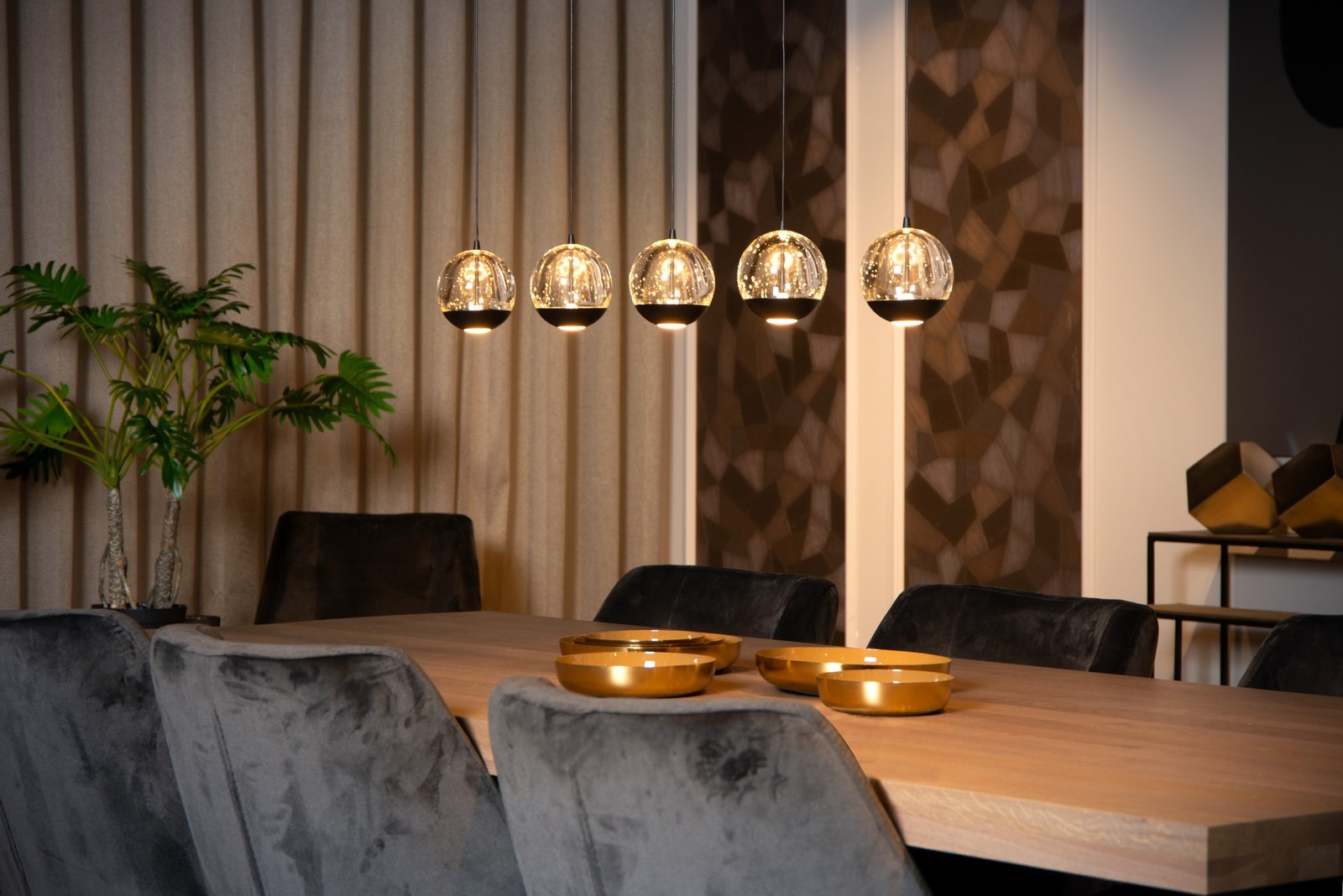 Nowoczesna lampa wisząca z pięcioma szklanymi kloszami nad drewnianym stołem w jadalni, z dekoracyjnymi złotymi miseczkami i rośliną doniczkową.