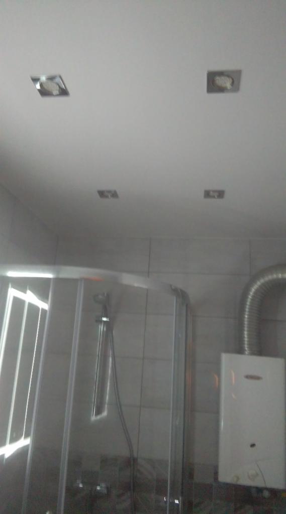 Wykończona łazienka z kabiną prysznicową z chromowanymi profilami i grzejnikiem przepływowym z odprowadzeniem spalin, widoczne płytki ścienne i cztery halogenowe lampy sufitowe.