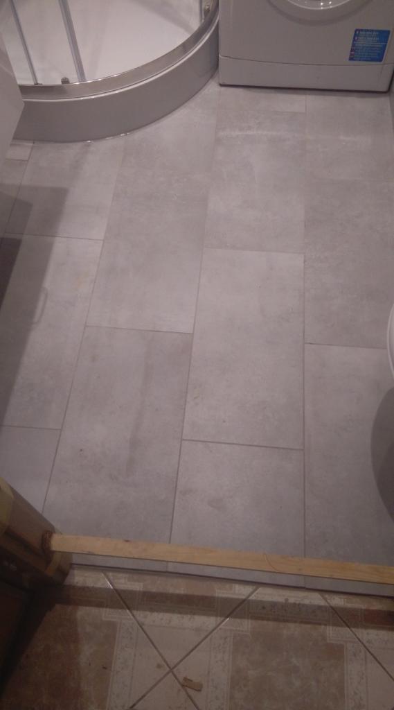 Szare płytki podłogowe w łazience, widoczna kabina prysznicowa i pralka, dolna część kadru pokazuje fragment starej podłogi z brązowymi płytkami.