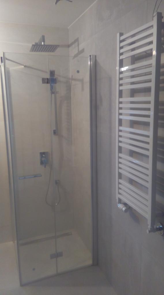 Nowoczesna kabina prysznicowa ze szklanymi drzwiami, deszczownicą i grzejnikiem drabinkowym w wyremontowanej łazience z jasnymi płytkami.