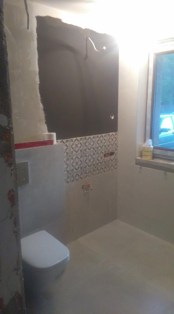 Niedokończona łazienka z częściowo położonymi płytkami dekoracyjnymi, widoczną instalacją elektryczną, wiszącą toaletą i oknem z widokiem na zewnątrz.