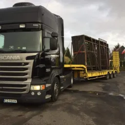 Czarno-srebrna ciężar&oacute;wka Scania R480 z ż&oacute;łtą naczepą niskopodwoziową przewożącą drewniane konstrukcje zabezpieczone czerwonymi pasami, na tle wiejskiego krajobrazu z budynkiem i polską flagą.