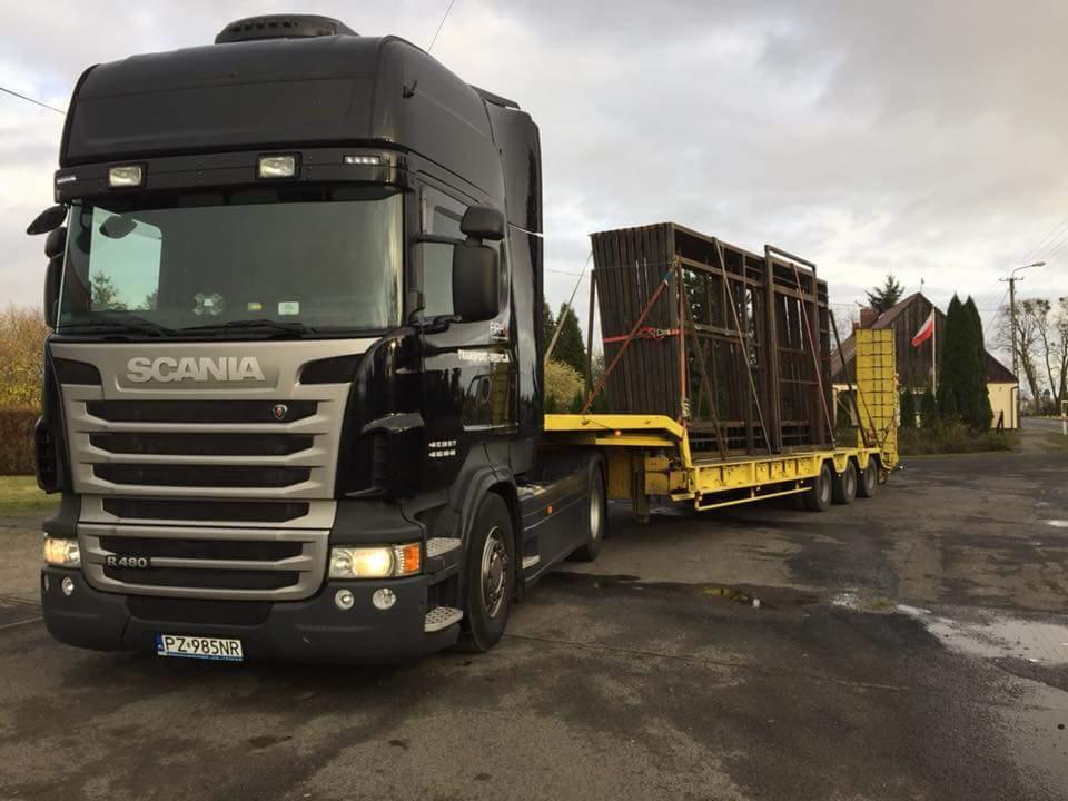 Czarno-srebrna ciężarówka Scania R480 z żółtą naczepą niskopodwoziową przewożącą drewniane konstrukcje zabezpieczone czerwonymi pasami, na tle wiejskiego krajobrazu z budynkiem i polską flagą.