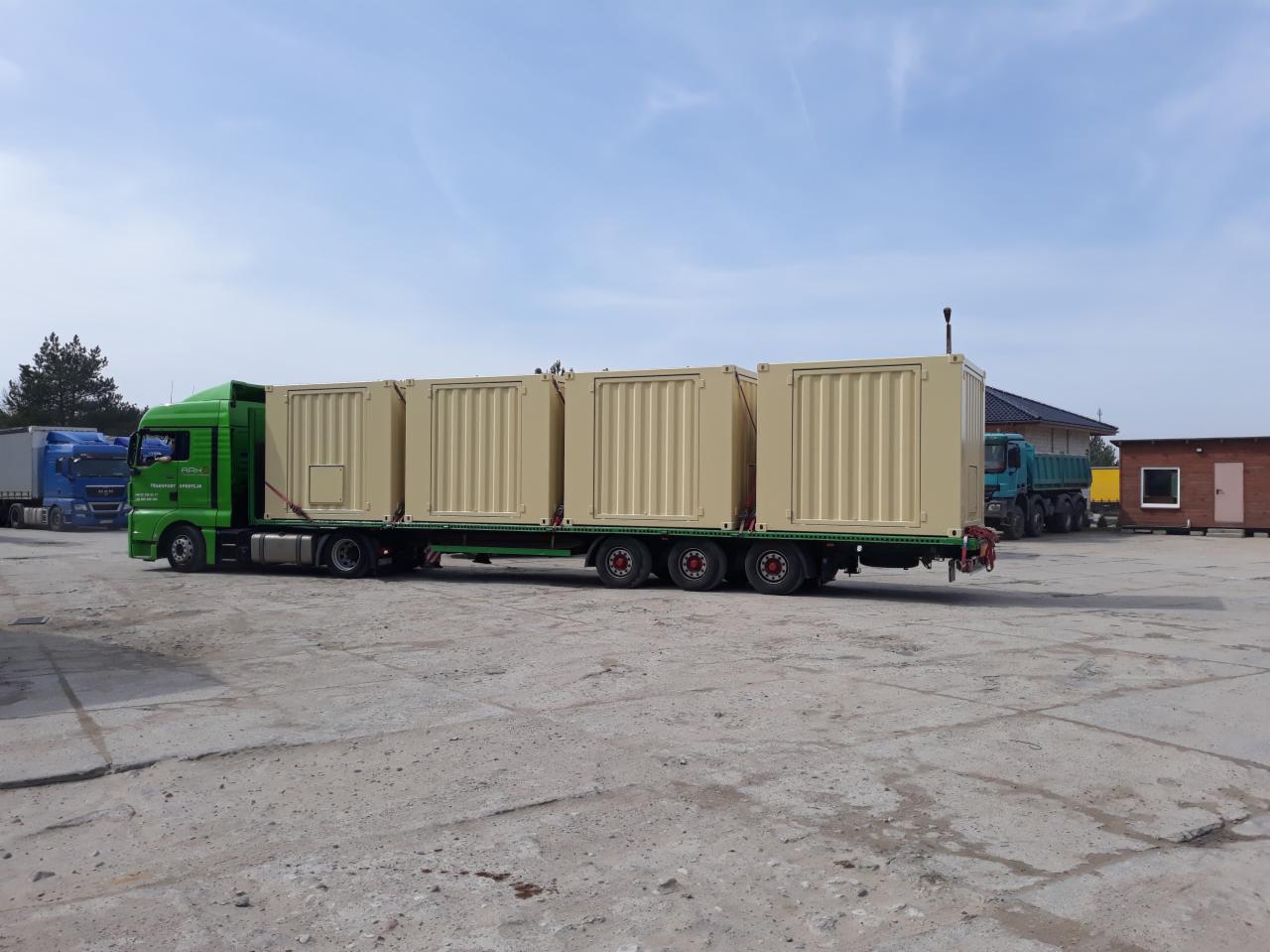 Zielona ciężarówka z naczepą przewożąca cztery beżowe kontenery o prążkowanej strukturze na placu manewrowym, widoczne inne pojazdy i budynki w tle.