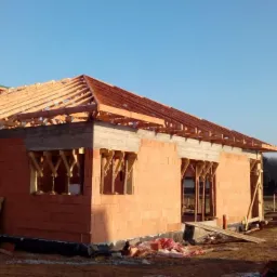 Surowy stan domu z czerwonej cegły, widoczna konstrukcja dachu z drewnianych belek, częściowo oszalowana, w tle błękitne niebo i drzewa.