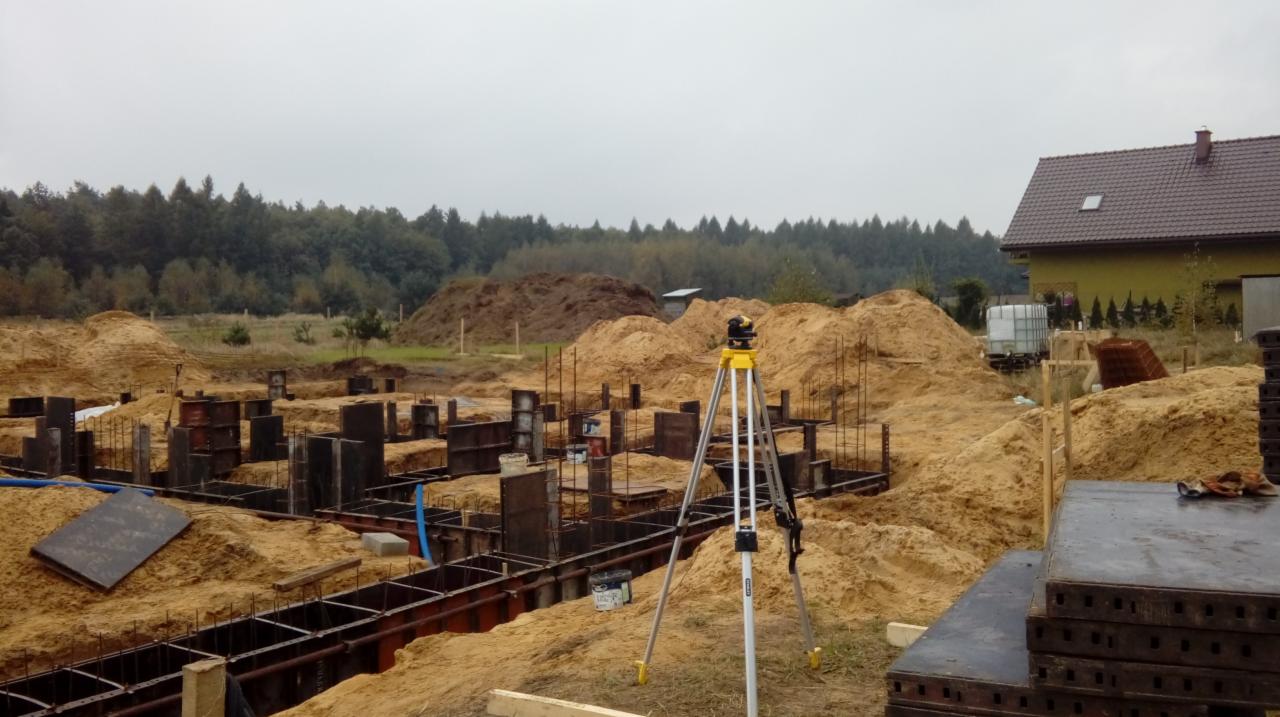 Fundamenty domu jednorodzinnego w trakcie budowy, widoczne szalunki, zbrojenia, piasek oraz niwelator laserowy na statywie, w tle dom i las.