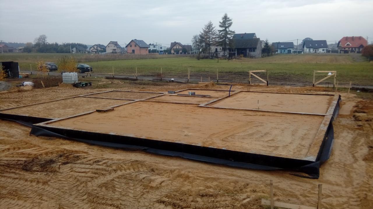 Przygotowany fundament pod budowę domu jednorodzinnego, widoczne drewniane szalunki, podsypka piaskowa, czarna folia hydroizolacyjna, teren wyrównany, w tle zabudowa jednorodzinna i drzewa.