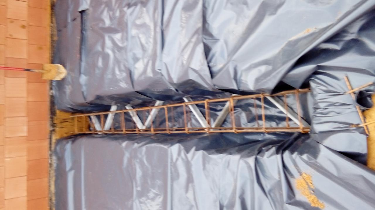 Zbrojenie fundamentu w wykopie zabezpieczonym folią, widoczna ceglana ściana i łopata oparta o nią.