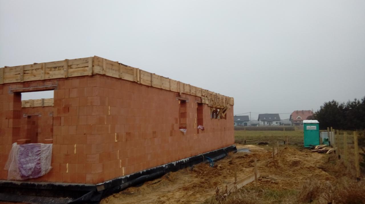 Surowy stan domu murowanego z czerwonej cegły, widoczne drewniane szalunki nad otworami okiennymi i drzwiami, w tle zarys zabudowań mieszkalnych i przenośna toaleta.