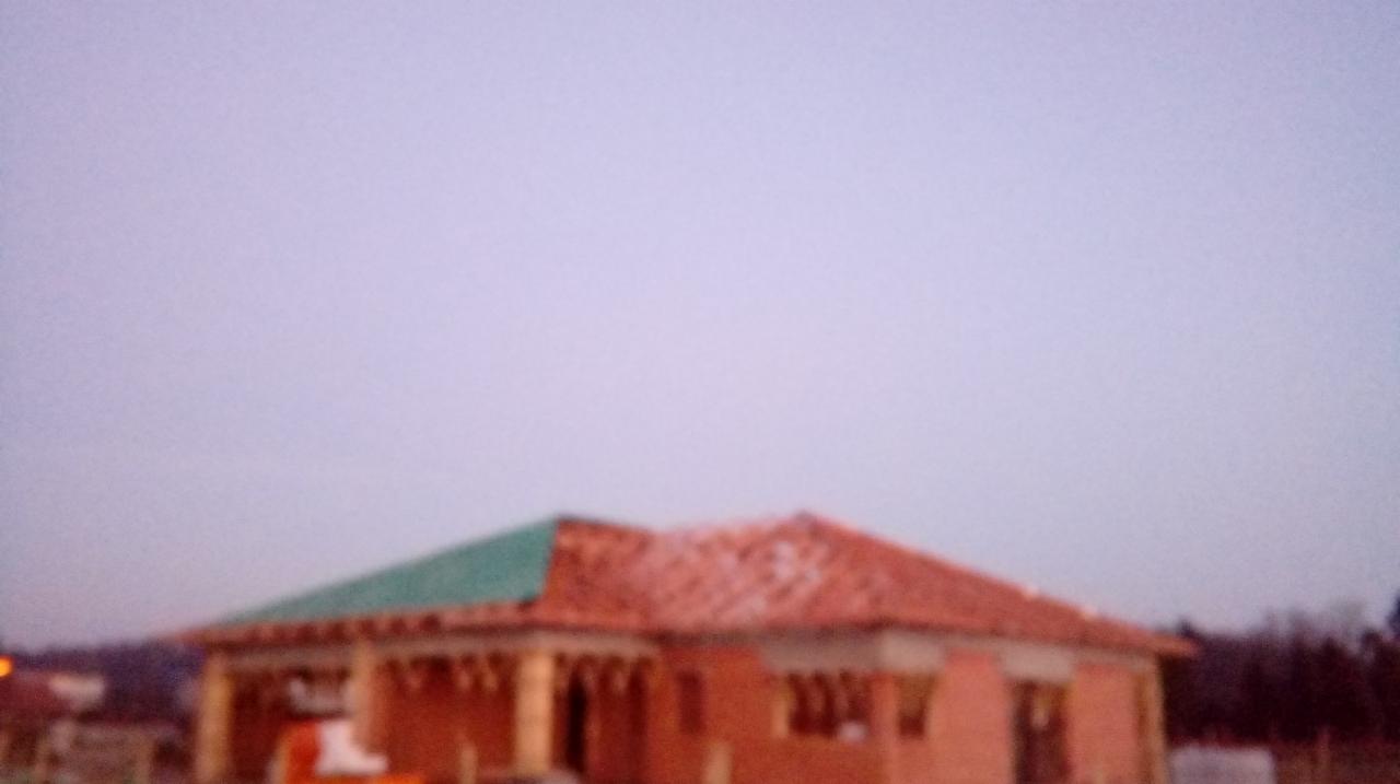 Niedokończony dom jednorodzinny z czerwoną dachówką, częściowo pokryty zieloną folią ochronną, widoczny w pochmurny dzień.