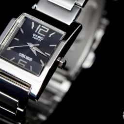 Zdjęcie zbliżeniowe kwadratowego zegarka Casio Quartz z metalową bransoletą na czarnym tle, z widocznym logo studia fotograficznego w prawym dolnym rogu.