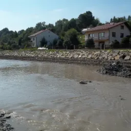 Brzeg rzeki umocniony kamieniami i betonem, w tle zabudowa mieszkalna i zieleń. Ujęcie z poziomu wody, woda mętna, prąd rzeki widoczny.