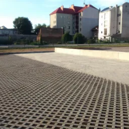 Ułożona szara kostka ażurowa na parkingu miejskim w słoneczny dzień. Część parkingu wyłożona gładką kostką. W tle budynki mieszkalne i drzewa.