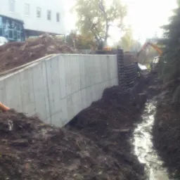 Betonowy mur oporowy w ziemi, z widocznym wykopem i maszynami budowlanymi w tle. Świeżo ułożone rury na pierwszym planie, budynek w oddali.