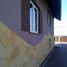 Elewacja domu z dekoracyjnym wykończeniem kamiennym na dole i drewnianymi okiennicami, widok z boku na kostkę brukową i ogrodzenie w tle.