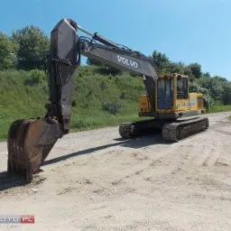 Żółta koparka gąsienicowa Volvo z łyżką zagłębioną w ziemi na placu budowy, widok z boku, w tle zielony nasyp i drzewa.