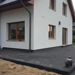 Nowoczesny dom z szarą kostką brukową na podjeździe i tarasie, widoczne niedokończone fragmenty i stos kostki, piasek w tle.