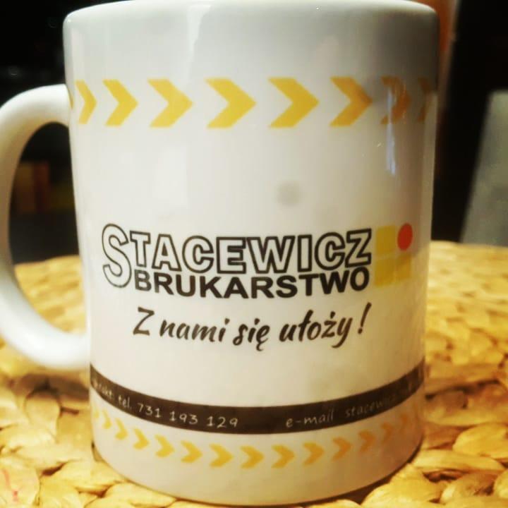 Biały kubek z logo firmy brukarskiej Stacewicz, z hasłem 'Z nami się ułoży!', numerem telefonu i adresem e-mail, ustawiony na plecionym podkładce.