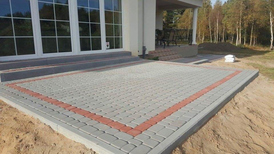Nowo ułożony taras z szarej kostki brukowej z obramowaniem z czerwonej kostki, przylegający do domu z dużymi oknami i schodami, z widocznymi workami z materiałem budowlanym w tle.