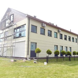 Damian Hołojda Firma Handlowo-Usługowa Profibud - Dwukondygnacyjny budynek w trakcie prac elewacyjnych. Na rusztowaniu dwóch pracowników. Widoczne farby, narzędzia i świeżo pomalowana część elewacji.