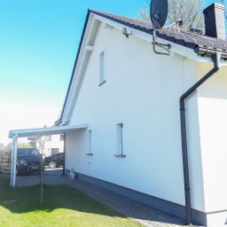 Damian Hołojda Firma Handlowo-Usługowa Profibud - Boczny widok domu po tynkowaniu elewacji na biało, z widocznym fragmentem dachu, rynny i podjazdu z zaparkowanym samochodem w słoneczny dzień.