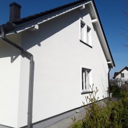 Damian Hołojda Firma Handlowo-Usługowa Profibud - Boczny widok domu po tynkowaniu elewacji w Goczałkowicach-Zdroju. Biała fasada, ciemna dachówka, okna z białymi ramami. Widoczne rynny i fragment ogrodu.