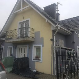 Damian Hołojda Firma Handlowo-Usługowa Profibud - Malowanie elewacji domu jednorodzinnego na szaro. Widoczne rusztowania, drabiny i częściowo pomalowane ściany. Prace w toku, przygotowanie do malowania.