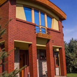 Damian Hołojda Firma Handlowo-Usługowa Profibud - Dwupiętrowy dom z czerwonej cegły z drewnianymi elementami, balkonem i numerem 22, widziany z dołu pod kątem, z fragmentem drzewa iglastego w kadrze.