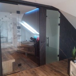 Damian Hołojda Firma Handlowo-Usługowa Profibud - Szklane drzwi przesuwne z czarnymi okuciami, oddzielające łazienkę z wanną od reszty pomieszczenia. Widoczne drewniane podłogi i skośny sufit.