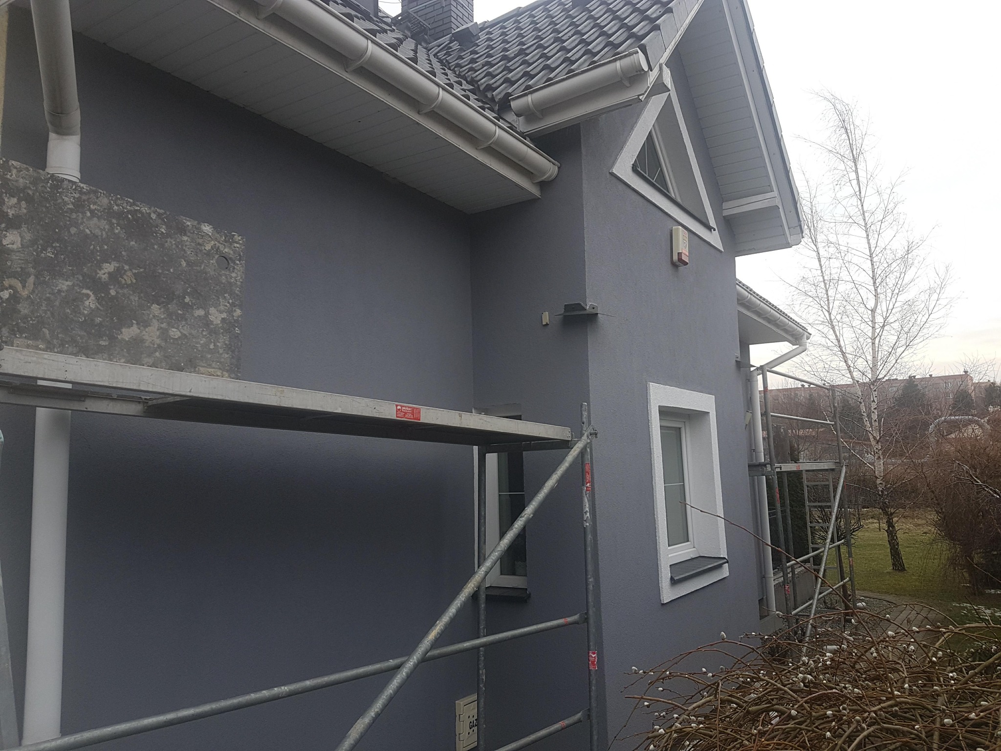 Szara elewacja domu po renowacji, widoczna z rusztowania. Świeżo pomalowana ściana, białe okna i rynny. W tle drzewa bez liści i fragmenty innych budynków.