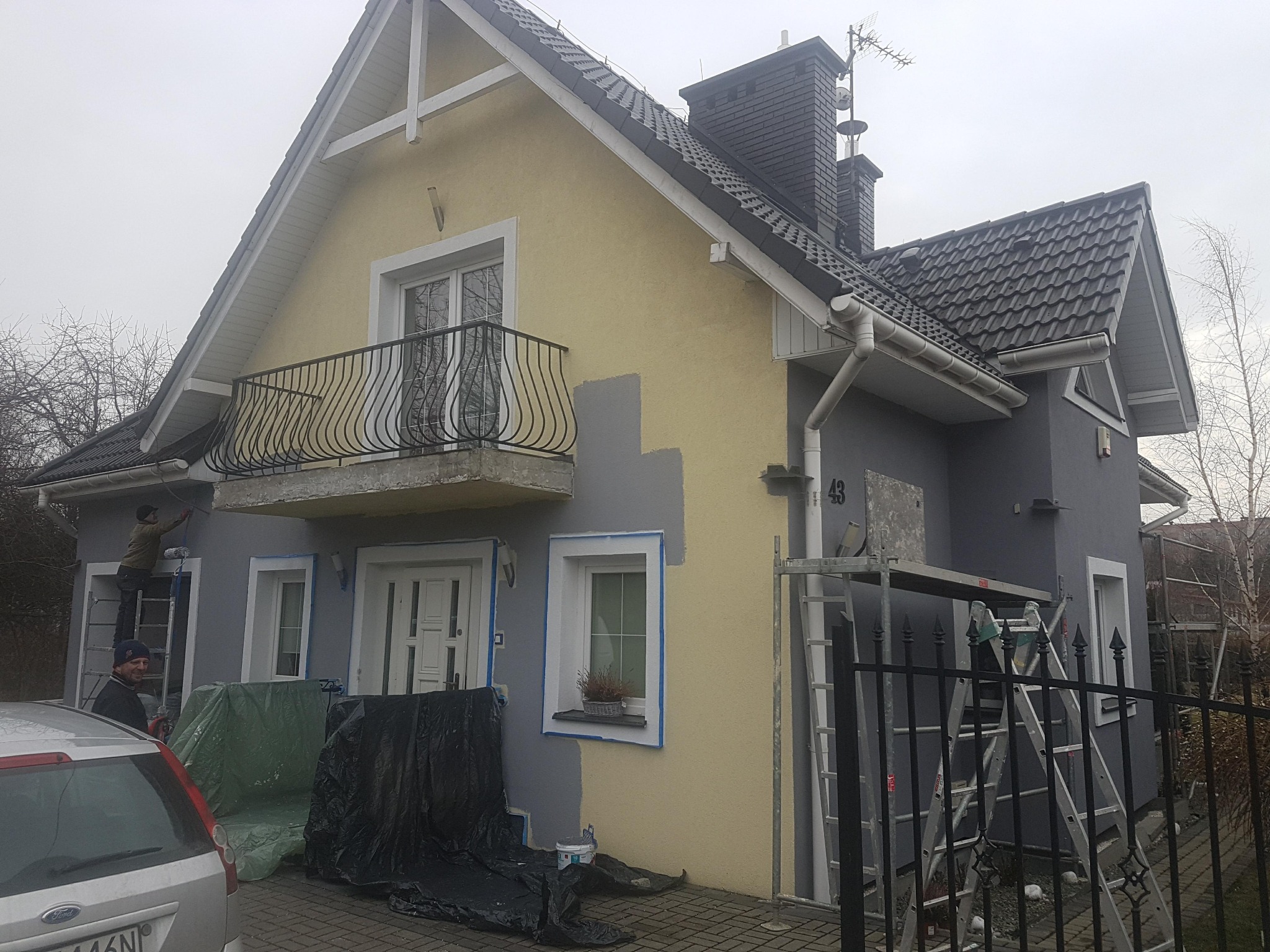 Malowanie elewacji domu jednorodzinnego na szaro. Widoczne rusztowania, drabiny i częściowo pomalowane ściany. Prace w toku, przygotowanie do malowania.