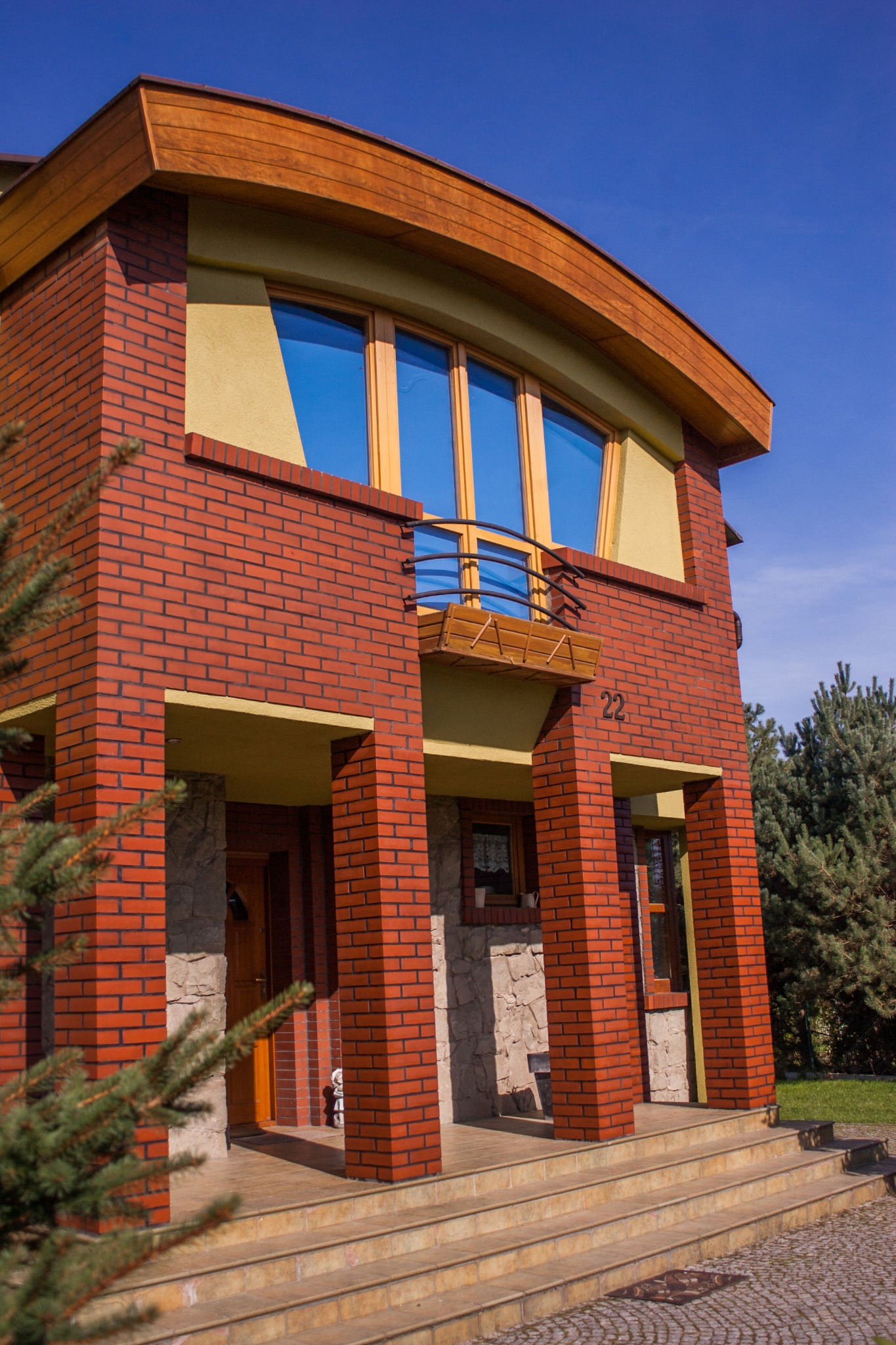 Dwupiętrowy dom z czerwonej cegły z drewnianymi elementami, balkonem i numerem 22, widziany z dołu pod kątem, z fragmentem drzewa iglastego w kadrze.