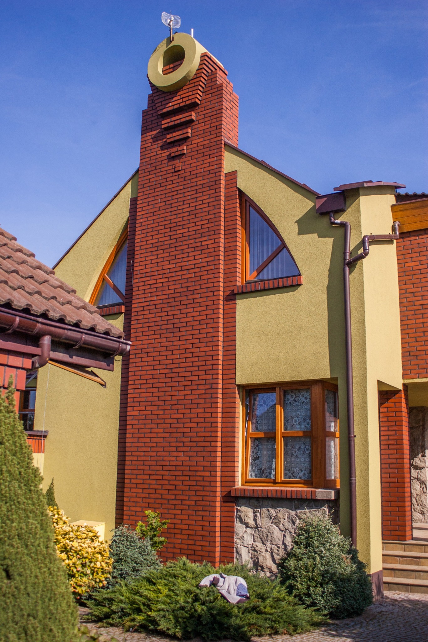 Dwupiętrowy dom z ceglanym kominem zakończonym okręgiem, żółtymi ścianami i trójkątnym oknem. Widoczne rynny i fragment dachu z dachówką.