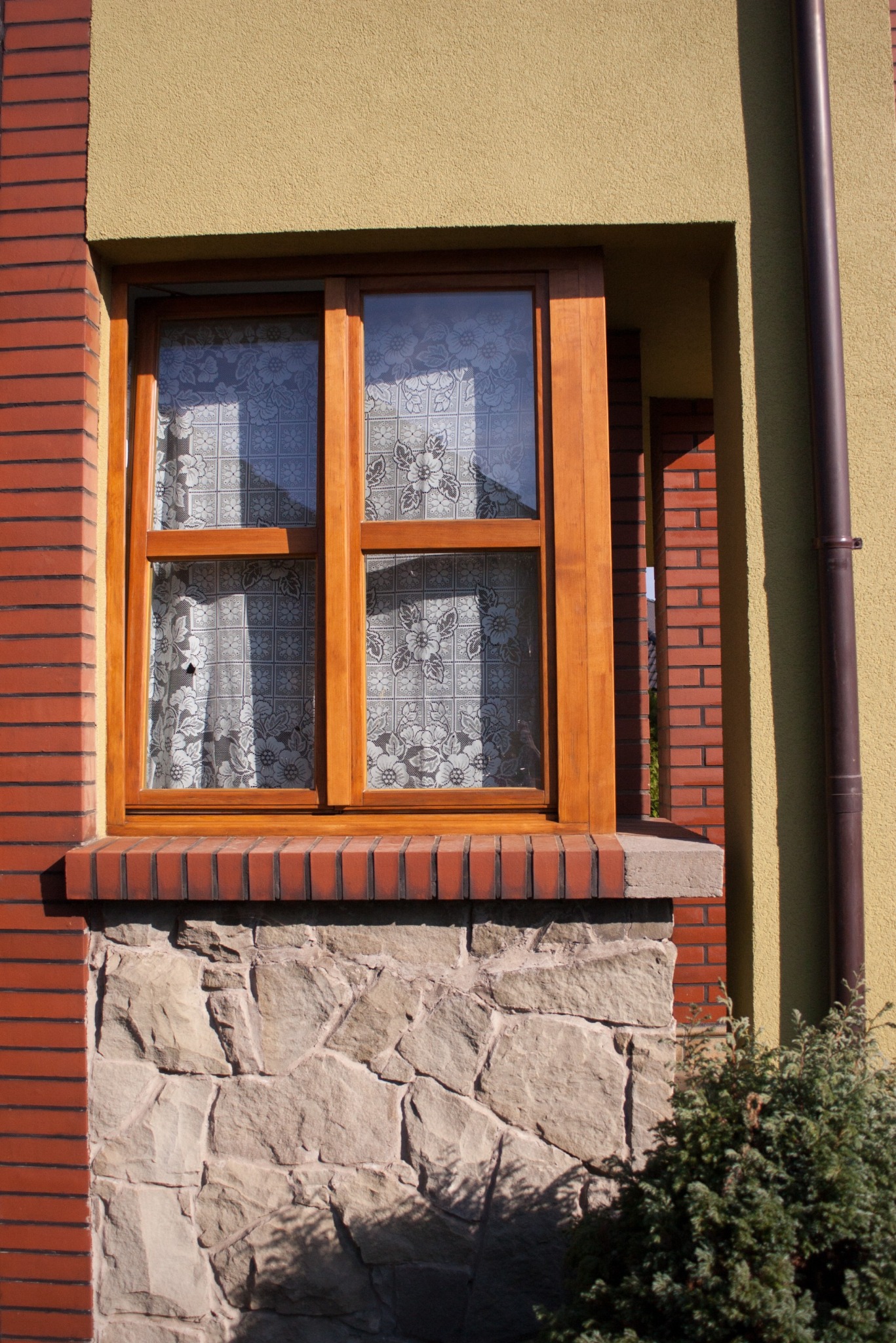 Drewniane okno z firanką w elewacji budynku z żółtym tynkiem i czerwoną cegłą, osadzone na kamiennej podmurówce. Widoczny fragment rynny i krzewu.