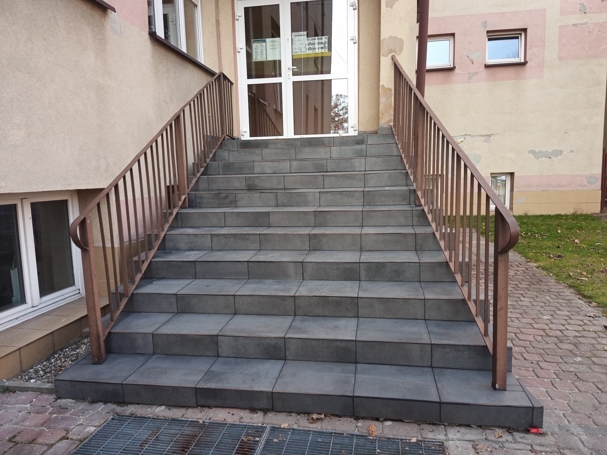 Zewnętrzne schody wejściowe wyłożone szarą, kwadratową płytką, z metalową balustradą w kolorze brązowym. Widoczne drzwi wejściowe i fragment elewacji budynku.