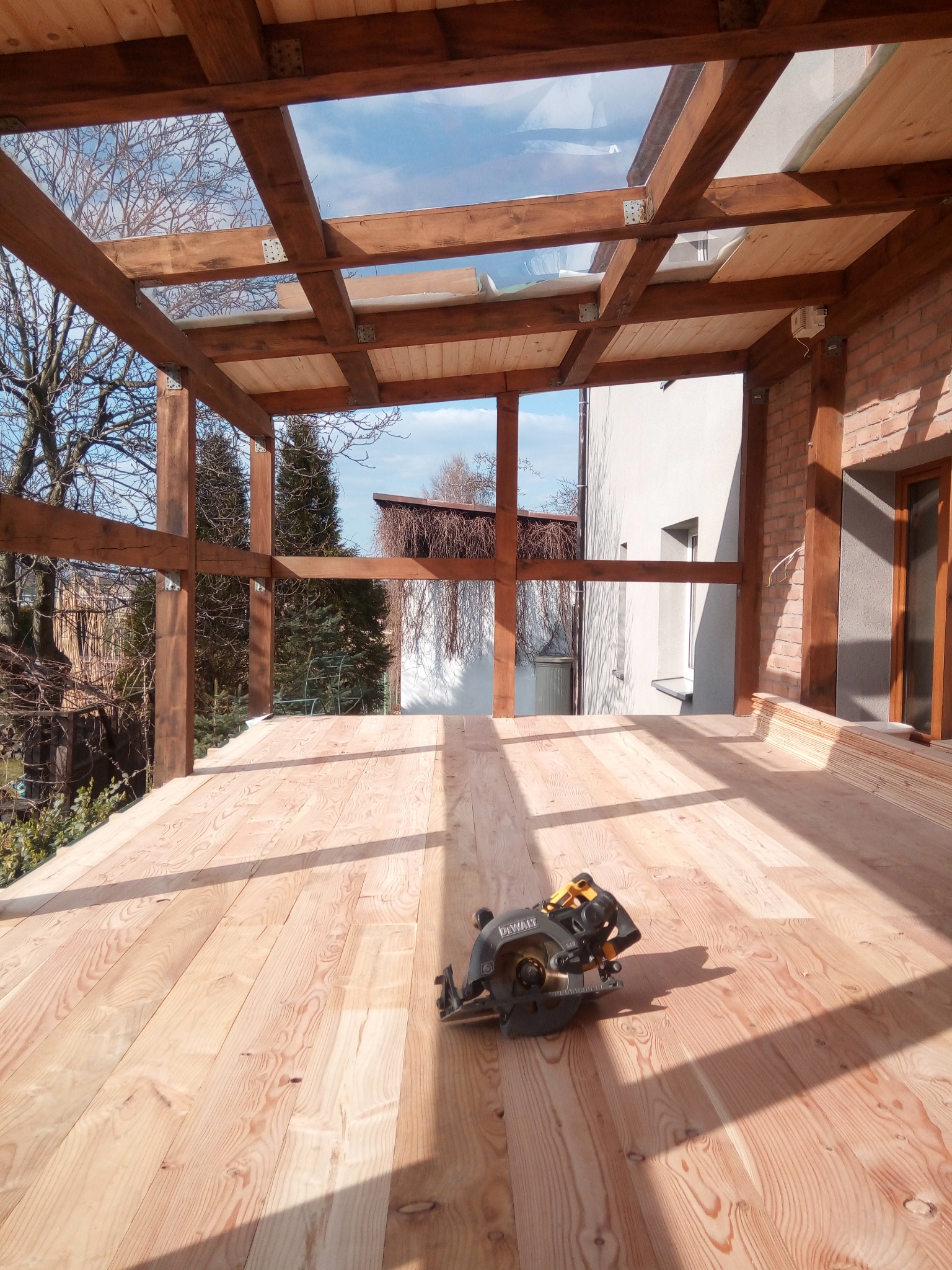 Drewniana konstrukcja z przeszklonym dachem w trakcie budowy. Na podłodze leży piła tarczowa. Widoczne elementy konstrukcyjne i naturalne światło.