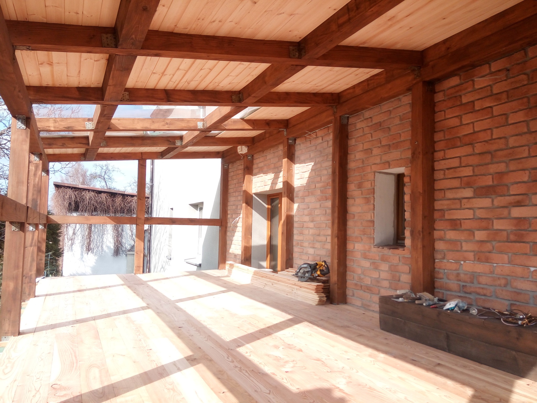 Zadaszony taras z drewnianą konstrukcją i ceglaną ścianą. Widoczne narzędzia i materiały budowlane, co sugeruje etap wykończeniowy. Jasne drewno podłogi i belek.