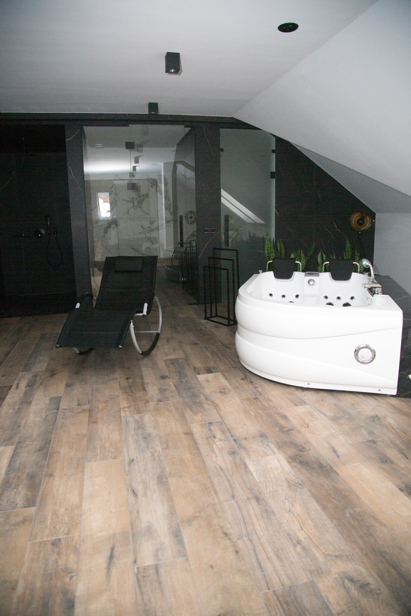 Nowoczesna łazienka na poddaszu z drewnopodobną podłogą, białą wanną z hydromasażem i czarną kabiną prysznicową. Widoczny leżak relaksacyjny.