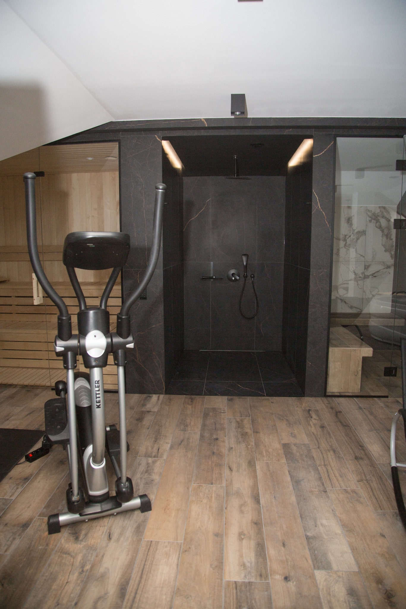 Nowoczesna łazienka z sauną i kabiną prysznicową wykończoną czarnym marmurem, drewniana podłoga, sprzęt do ćwiczeń. Minimalistyczny design.